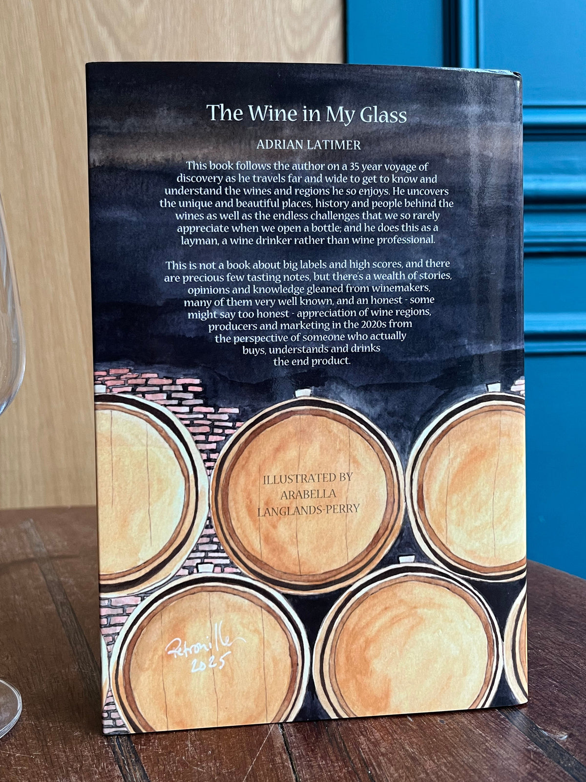 Book titled 'The Wine in My Glass' by Adrian Latimer on a wooden surface with a glass of wine.