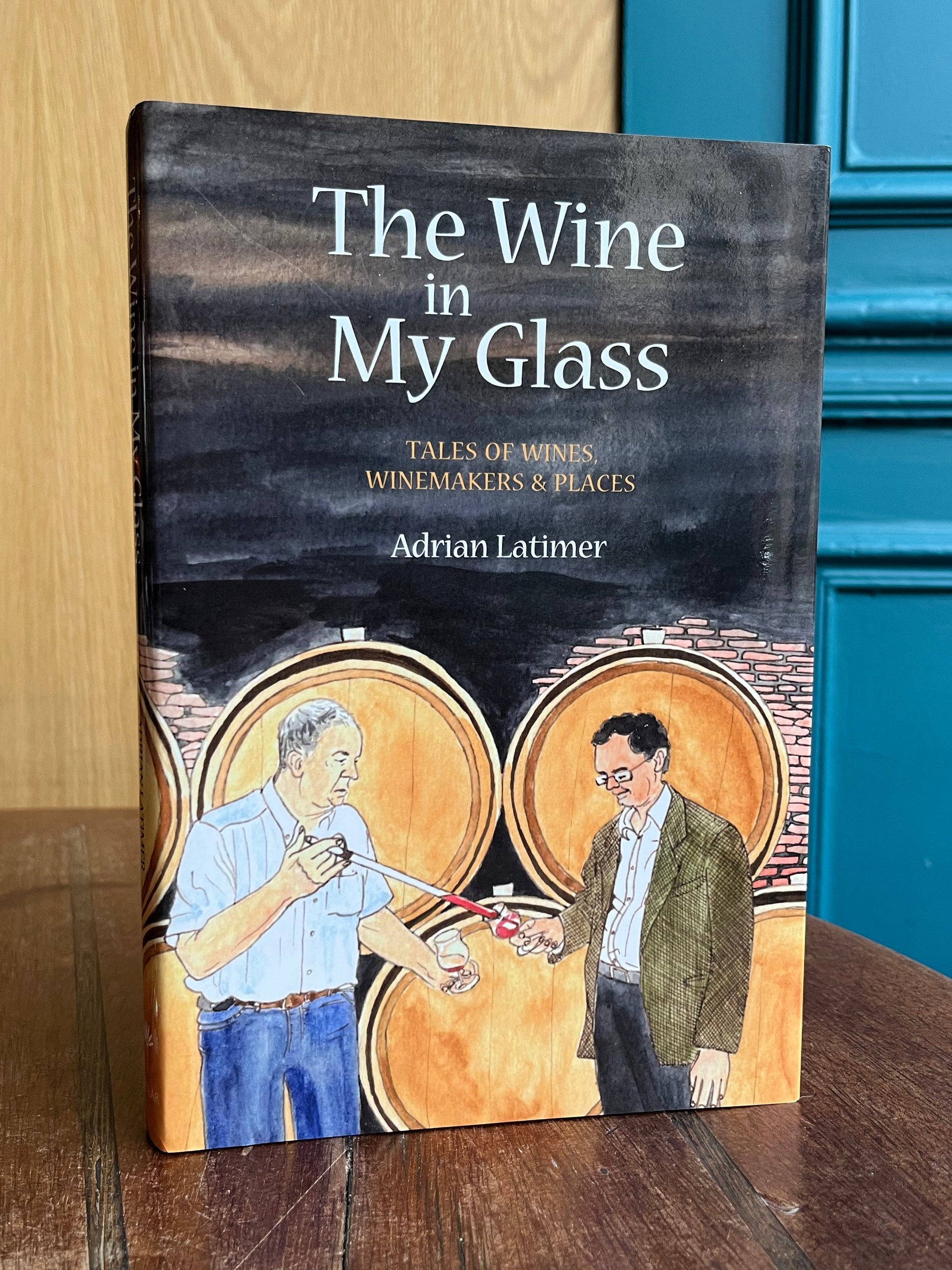 Book titled 'The Wine in My Glass' by Adrian Latimer on a wooden surface with a glass of wine.